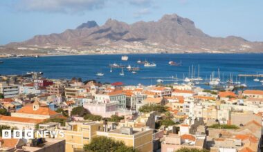 Elena Walsh, a 64-year-old woman with short ginger hair and wearing a red shirt, smiles with her arms around her husband, a main with grey hair and a blue shirt, and her son, a younger man with short brown hair, a beard and a grey jumper.
