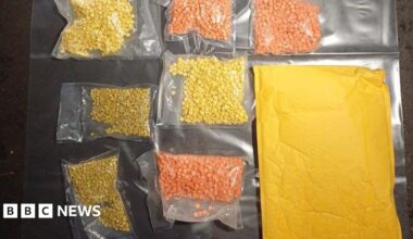Clear plastic bags containing hundreds of yellow and orange pills are laid out on a plastic sheet. A brown envelope is lying next to the bags.