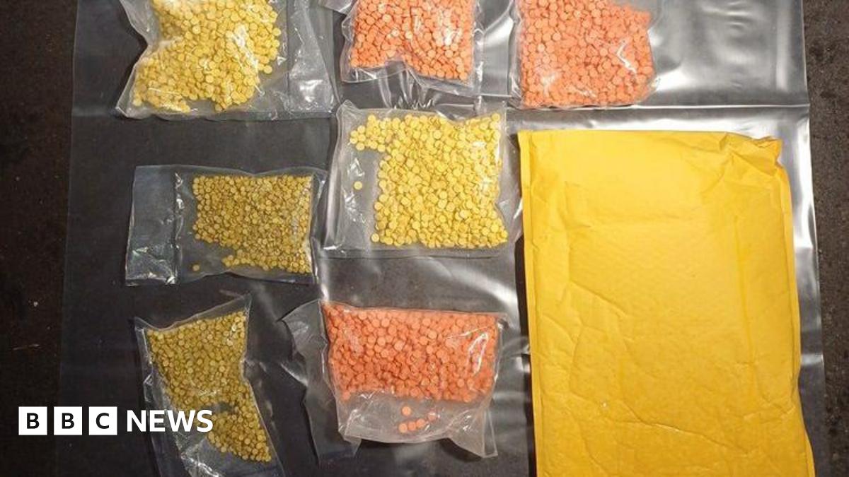 Clear plastic bags containing hundreds of yellow and orange pills are laid out on a plastic sheet. A brown envelope is lying next to the bags.