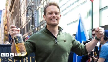 A man in a green top, smiling and holding a bottle of whisky in his hand, with a column of marchers behind him