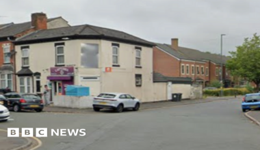 General view of Montgomery Street in Birmingham. Screenshot shows the junction of a road, on the corner there is a white two-storey building with a purple sign. On the right of the image there is a wall next to the rear view of a painted white brick building. On the side is a white sign with the words Hi-ton in blue