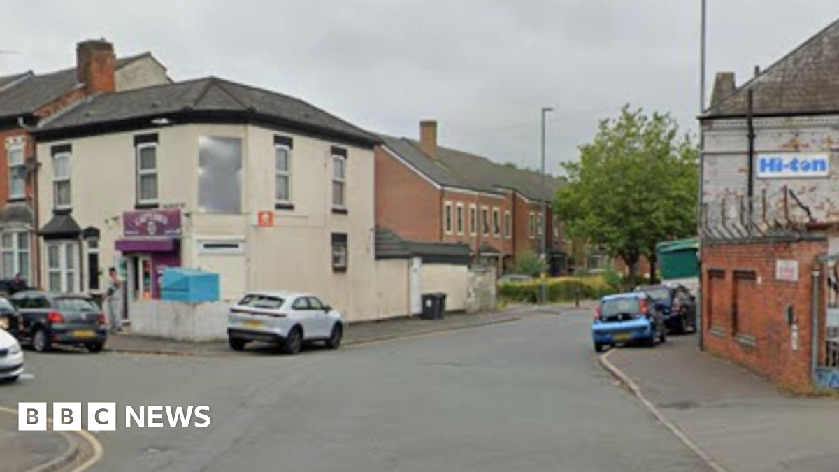 General view of Montgomery Street in Birmingham. Screenshot shows the junction of a road, on the corner there is a white two-storey building with a purple sign. On the right of the image there is a wall next to the rear view of a painted white brick building. On the side is a white sign with the words Hi-ton in blue