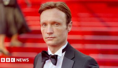 Hans Obma stood on a red carpet. Red stairs can be seen behind him, he wears a black suit jacket, white shirt and black bow tie. He has blue eyes and brown hair and looks side on down the lens.