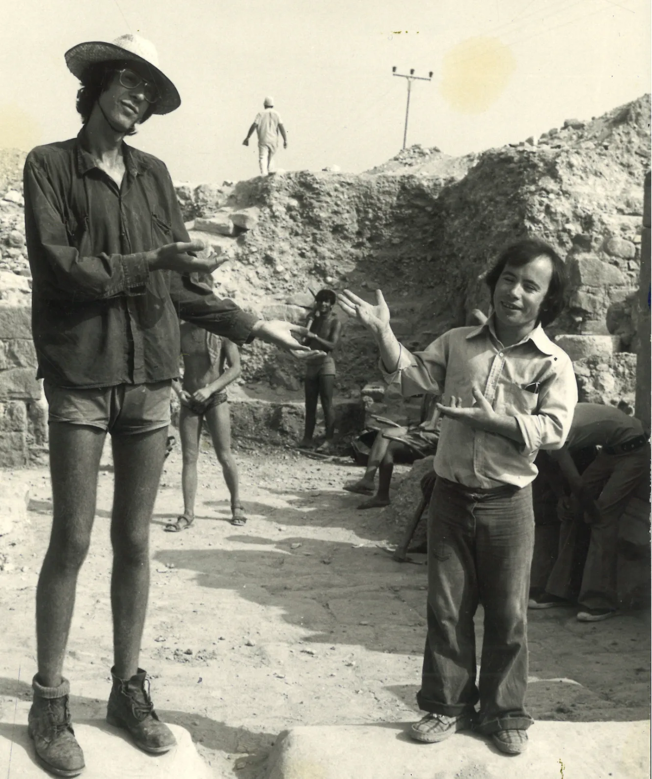 Robert Goodman, a tall man in a straw hat and sunglasses, stands with his palms up, facing a shorter man who gestures with open hands on an archaeological dig.