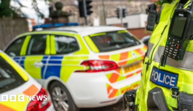 Close up of the Police badge on the front of a hi-vis jacket, with two police cars in the background