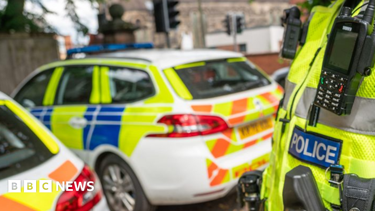 Close up of the Police badge on the front of a hi-vis jacket, with two police cars in the background