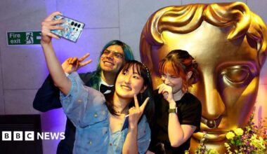 A group of three young people pose for a selfie in front of a large, golden Bafta mask statue. The young woman at the front of the group holds up her camera phone and throws up a peace sign, as does the young man with green hair to her right. A young woman to her left places the back of her hand against her mouth as she giggles.