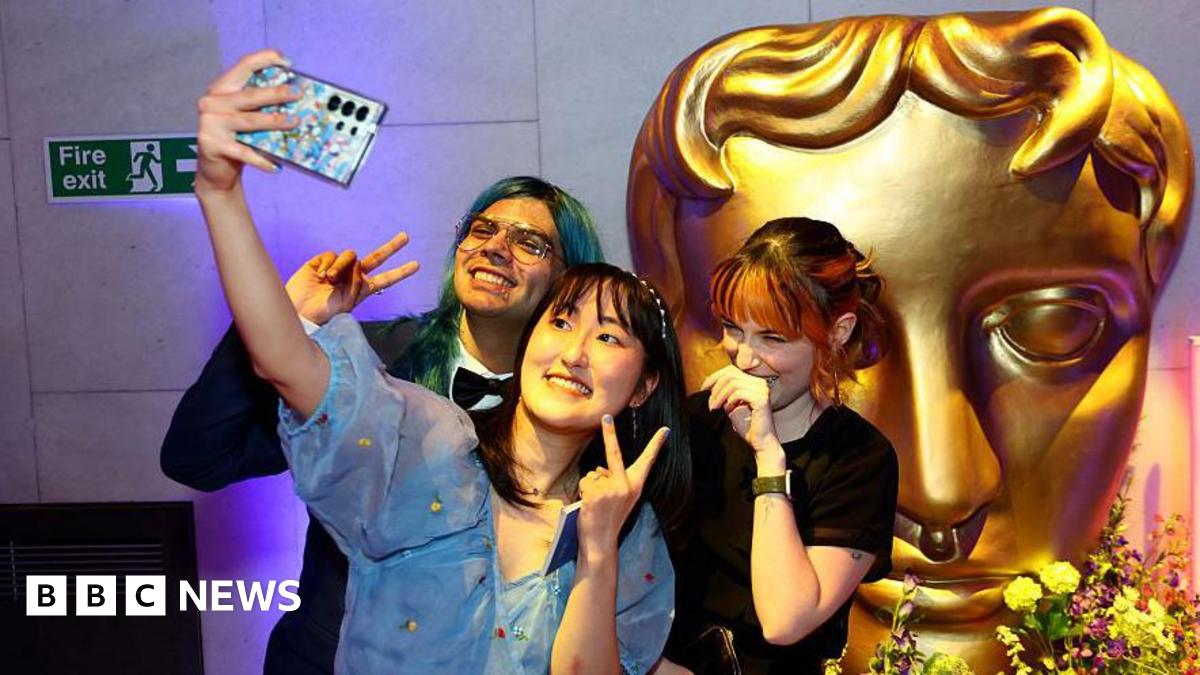 A group of three young people pose for a selfie in front of a large, golden Bafta mask statue. The young woman at the front of the group holds up her camera phone and throws up a peace sign, as does the young man with green hair to her right. A young woman to her left places the back of her hand against her mouth as she giggles.