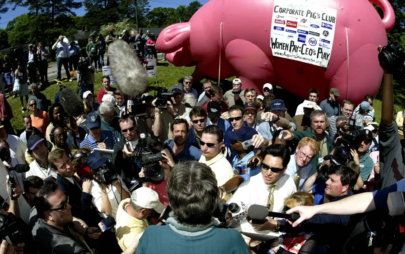 Martha Burk speaks to the media with an inflatable pink pig in the background, labeled "Corporate Pig's Club" and "Women Pay: CEOs Play".