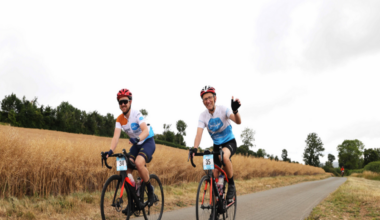 Two men cycling along a lane in a field on a charity bike ride. They are wearing blue and white lycra tops, black shorts, and red helmets. Their bikes are red and have a blue and white number card on the handlebars. The men are waving and smiling. It is a cloudy day but the grass on either side of the lane is scorched.