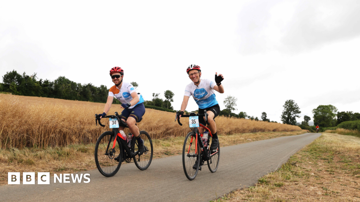 Two men cycling along a lane in a field on a charity bike ride. They are wearing blue and white lycra tops, black shorts, and red helmets. Their bikes are red and have a blue and white number card on the handlebars. The men are waving and smiling. It is a cloudy day but the grass on either side of the lane is scorched.
