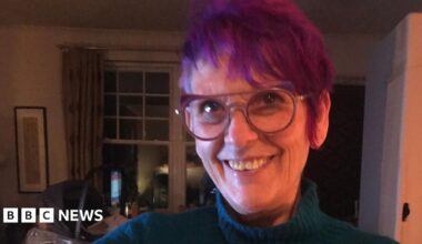 A woman with spiky purple hair and glasses, wearing a blue roll neck, smiles while stood inside a dimly-lit living room.