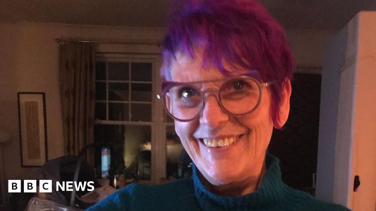 A woman with spiky purple hair and glasses, wearing a blue roll neck, smiles while stood inside a dimly-lit living room.