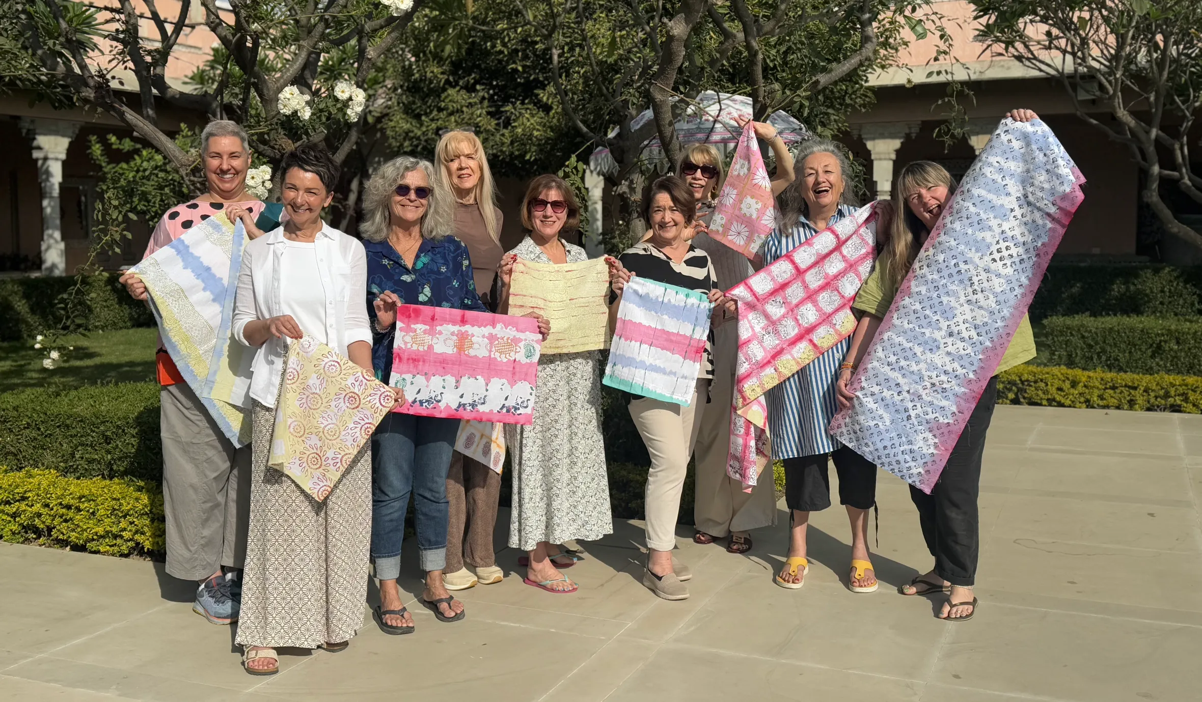 A group of ten women holding up patterned textiles in an outdoor garden.