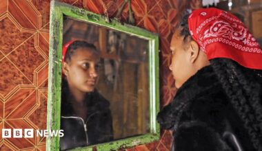 Nasibo wears a black hijab and is photographed with green plants behind her - she is looking away from the camera