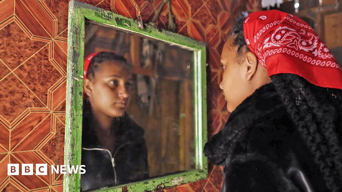 Nasibo wears a black hijab and is photographed with green plants behind her - she is looking away from the camera