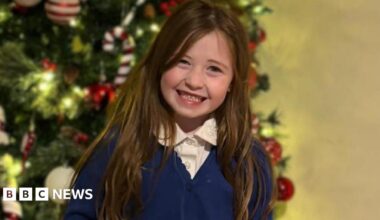 Nyla standing indoors in front of a white chest of drawers. She is smiling warmly at the camera with her hands held behind her back. She is wearing a school uniform consisting of a white collared shirt, a blue cardigan, a grey pleated skirt, grey tights, and black shoes. Her long, light brown hair falls neatly over her shoulders. On top of the chest of drawers behind her are decorative items, including a black vase with gold details and another vase containing dried foliage.