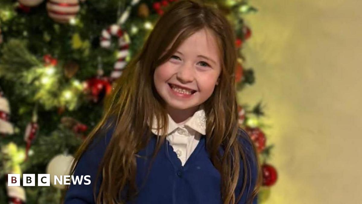Nyla standing indoors in front of a white chest of drawers. She is smiling warmly at the camera with her hands held behind her back. She is wearing a school uniform consisting of a white collared shirt, a blue cardigan, a grey pleated skirt, grey tights, and black shoes. Her long, light brown hair falls neatly over her shoulders. On top of the chest of drawers behind her are decorative items, including a black vase with gold details and another vase containing dried foliage.