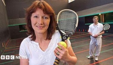 A woman with short red brown hair wearing a white t-shirt. She is standing on an indoor real tennis court which has colourful lines painted across the floor with numbers next to them. She is holding a small slim tennis-style racquet and a yellow tennis ball. There is a man standing in the background, also holding one of the racquets
