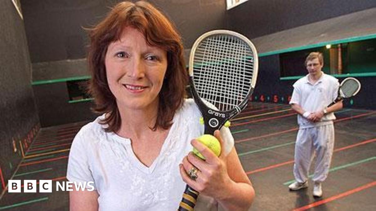 A woman with short red brown hair wearing a white t-shirt. She is standing on an indoor real tennis court which has colourful lines painted across the floor with numbers next to them. She is holding a small slim tennis-style racquet and a yellow tennis ball. There is a man standing in the background, also holding one of the racquets