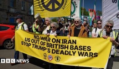 Protesters at the front of a demonstration are holding a large yellow anti-war banner, with many people following behind them.