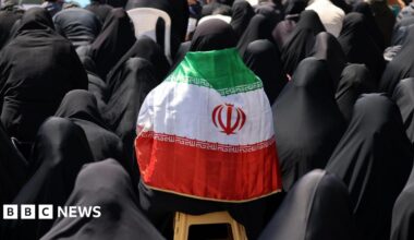 An Iranian woman wears a black hijab and chador and has a national flag draped over her shoulders as government supporters gather at Enghelab Square following the US-Iran ceasefire announcement, in Tehran, Iran (8 April 2026)
