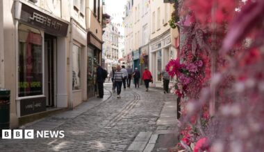 People walk along a street with shops on it in St Peter Port, Guernsey. The shop front closest to the camera has flowers outside. The street is cobbled.