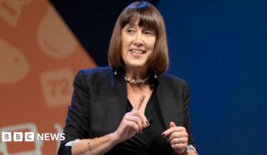 Jane Dodds speaking at a Liberal Democrat event, in front of a blue and orange background. She is wearing a black jacket and top.