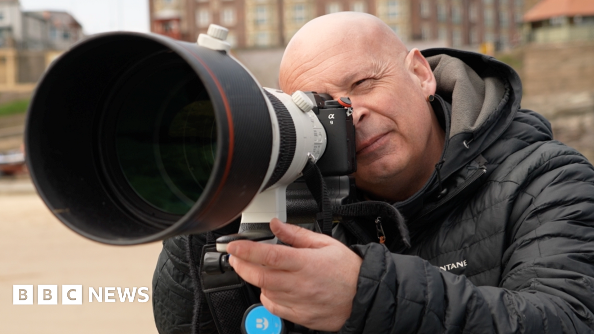 Owen Humphreys is holding his camera and taking a photo. The angle is such that it is like we are looking down the lens. Humphreys is looking off into the distance, picking his shot. He is bald and has a black earring in one ear. He is wearing a black puffer jacket. The coast wall behind him is out of focus.