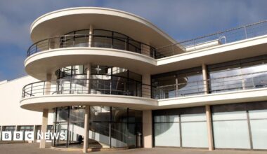 The De La Warr Pavilion’s modernist architecture has curved white balconies, with metal railings and a glass‑fronted stairwell with a spiral staircase inside. There are large rectangular windows looking out on to the open paved terrace in the foreground.