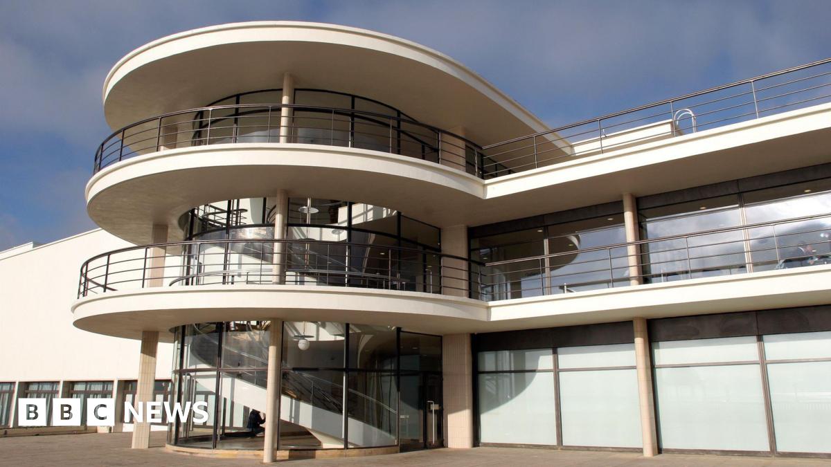 The De La Warr Pavilion’s modernist architecture has curved white balconies, with metal railings and a glass‑fronted stairwell with a spiral staircase inside. There are large rectangular windows looking out on to the open paved terrace in the foreground.