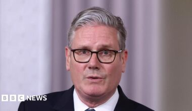 Nathan sitting in a pub with a drink. He is wearing a blue tracksuit top and has his arms crossed. He has a short beard and dark brown hair.