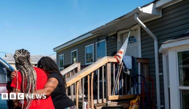 Women hug near the entrance of a home. There's a US flag there. Some damage can be seen to the roof