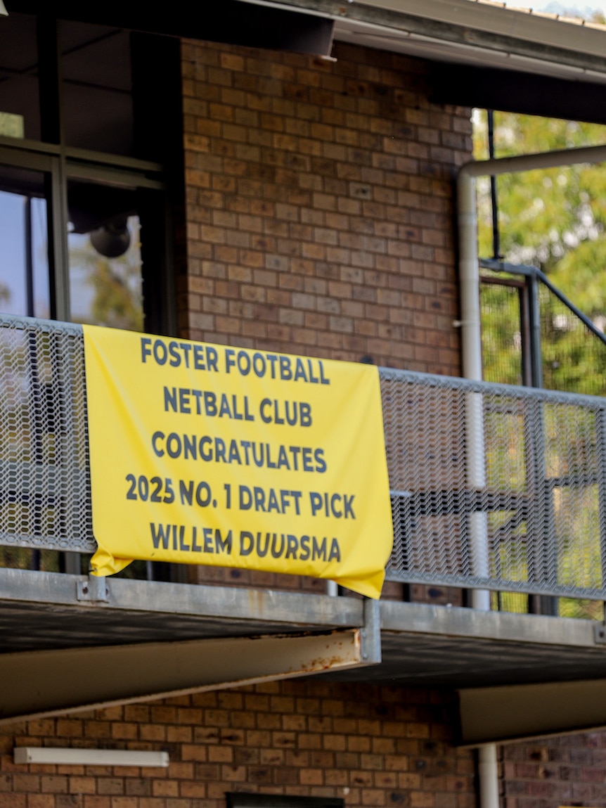 Yellow sign with text celebrating Willem Duursma hangs on balcony of brick building next to football oval.