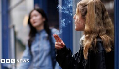 A young woman smokes outside a shop. She has long blonde hair and wears a leather jacket