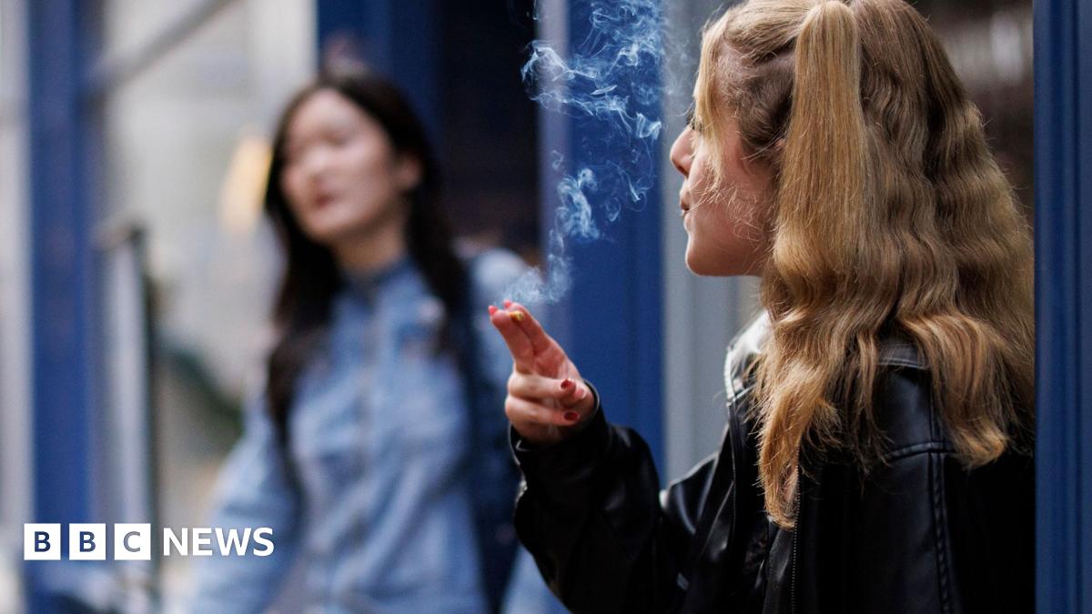 A young woman smokes outside a shop. She has long blonde hair and wears a leather jacket
