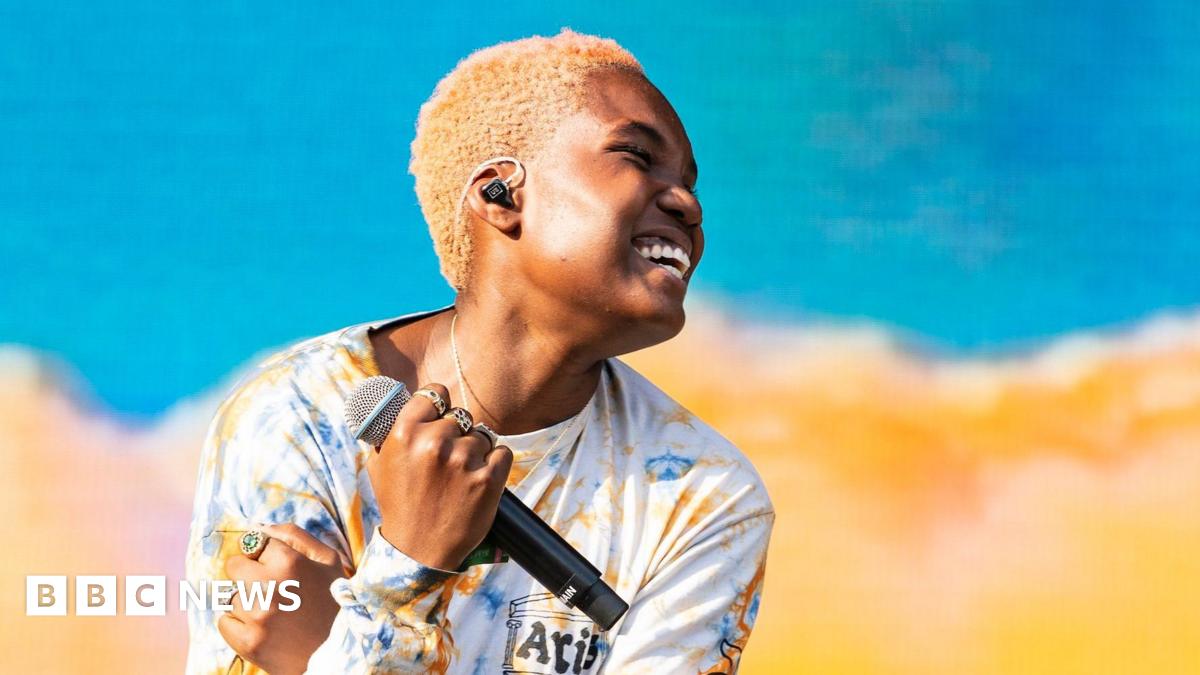 Arlo Parks gives a disarmingly carefree smile as she performs in Australia. She's holding a microphone in her right hand, and wearing a colourful long-sleeved t-shirt, whose blue and yellow colours are reflected on the stage behind her.