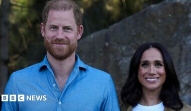 Harry stands next to Meghan. He is wearing a light blue shirt. They are both smiling.