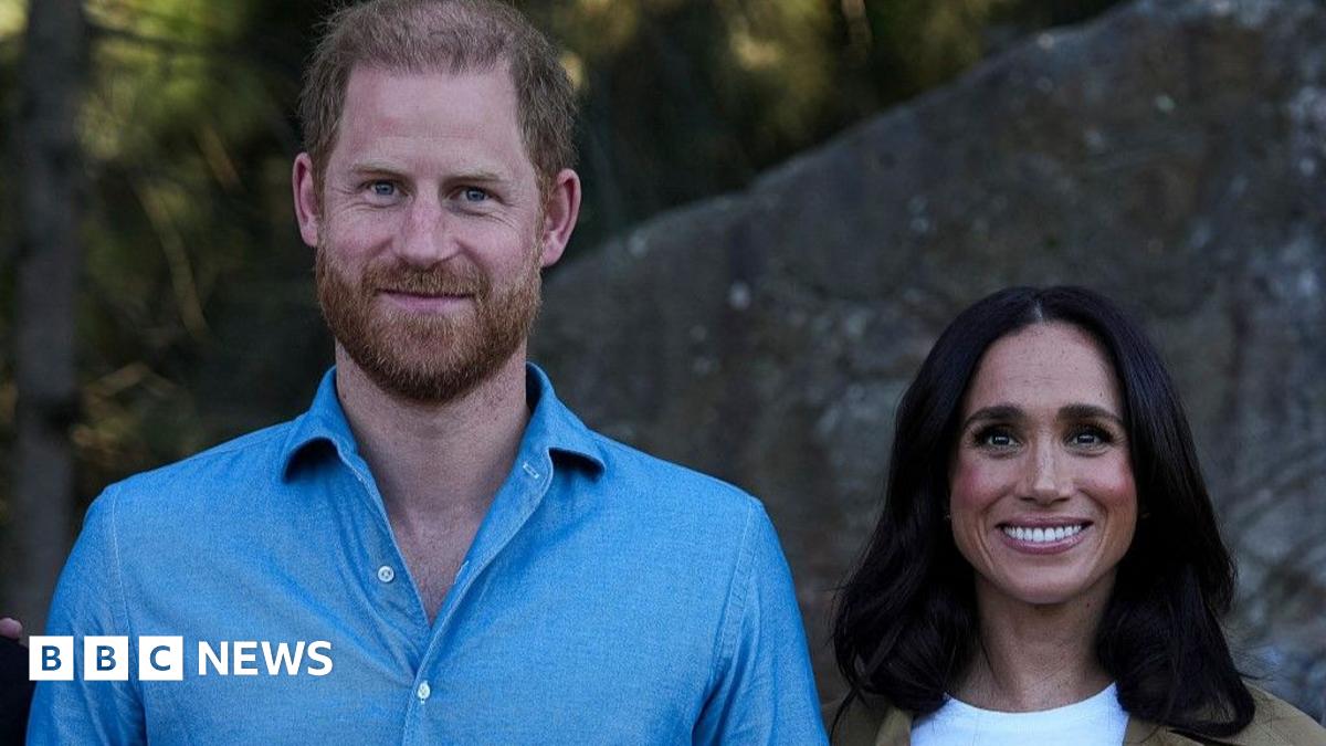 Harry stands next to Meghan. He is wearing a light blue shirt. They are both smiling.