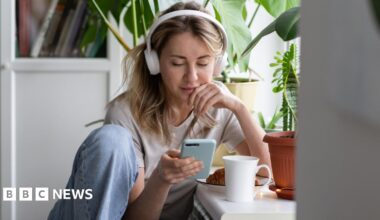 A stock shot of a young blonde woman wearing white headphone and looking at her phone.