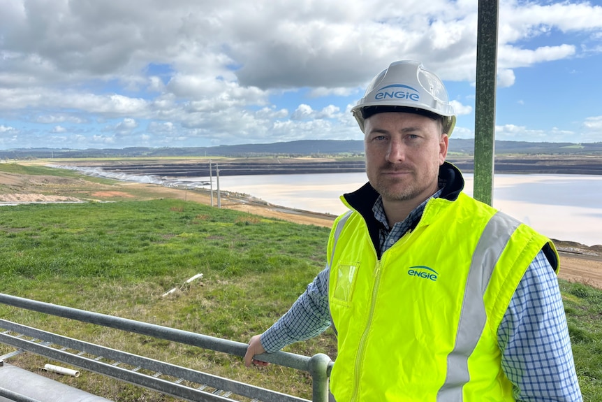 A young man with a brown beard wears safety gear and an engie helmer, standing in front of an empty mine pit