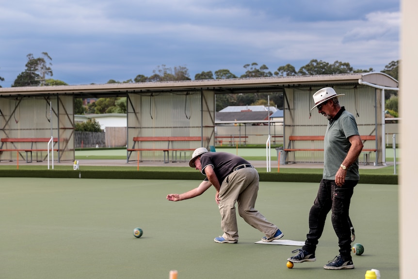 Two men stand on a bowling green, one is hunched over bowling, the other watches on.
