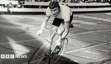 A black and white photo of a man cycling on a track looking strained as crowds cheer him on
