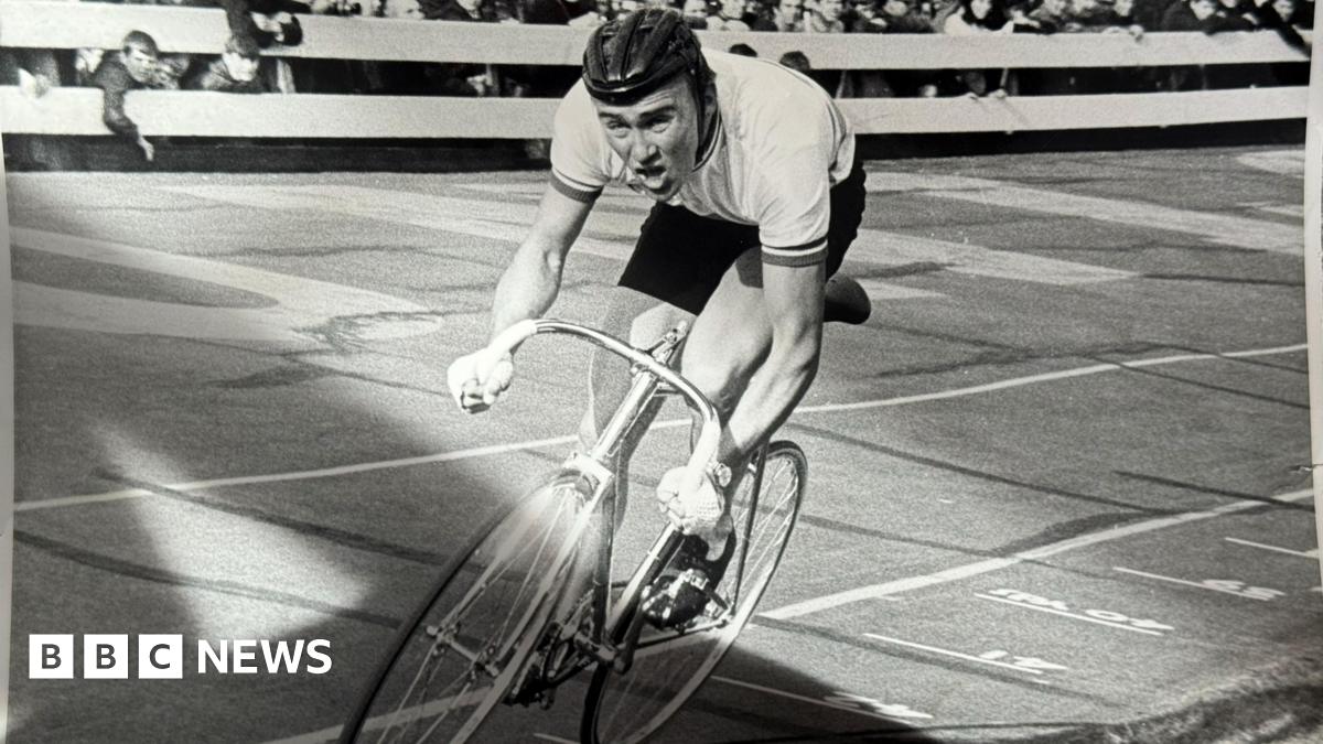 A black and white photo of a man cycling on a track looking strained as crowds cheer him on