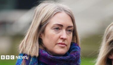 Esther Ghey, who has shoulder-length blonde hair, looks on as she attends an event. She has a blue and purple scarf around her neck.