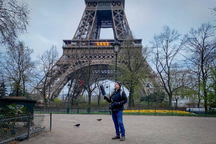 The researcher stands holding out a large black microphone, with the Eiffel Tower behind him.