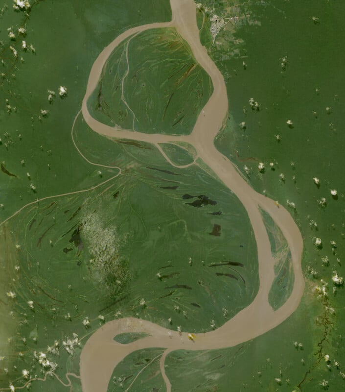 Aerial view of a winding river with muddy water flowing through a lush green landscape, surrounded by scattered white clouds and patches of vegetation.