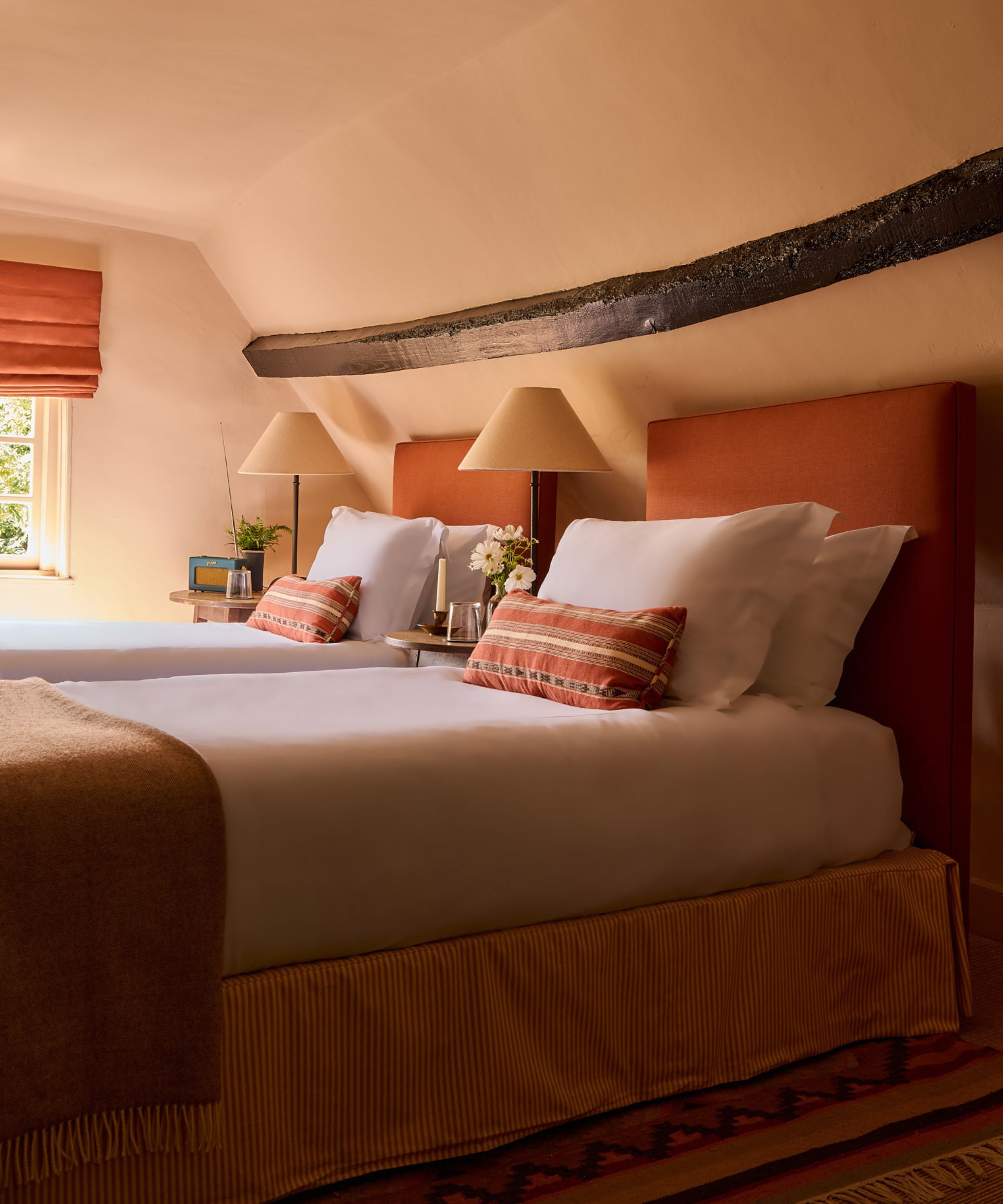 A loft bedroom with sloped ceilings color-drenched plaster pink, an exposed wood ceiling beam, and twin beds with terracotta headboards, crisp white bedding, and terracotta cushions and throws.