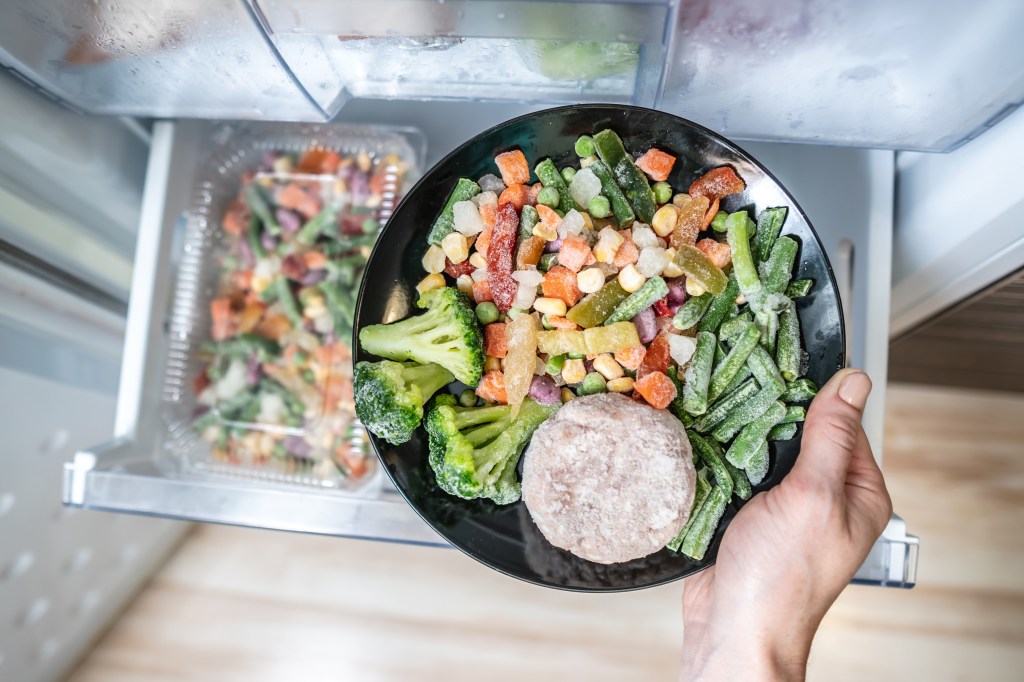 A hand holding a plate of frozen vegetables and a meat patty out of a freezer drawer.