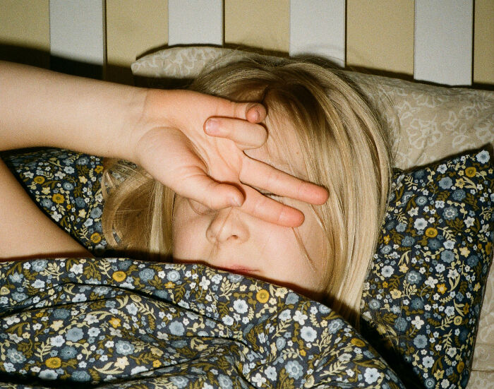 Healthcare worker resting in bed, partially covered with floral blanket, shielding eyes with hand in a cozy bedroom setting